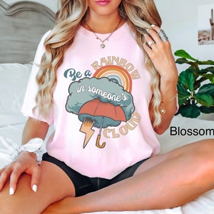 May include: A pink t-shirt with a graphic design of a rainbow in a cloud with an umbrella and lightning bolt. The text on the shirt reads "Be a rainbow in someone's cloud."