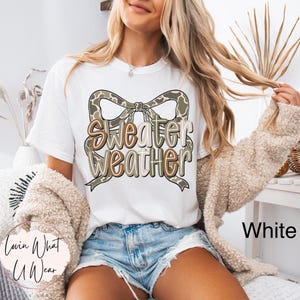 May include: White t-shirt with the text "Sweater Weather" in a bow design. The bow has a camouflage pattern. The shirt is paired with denim shorts and a beige cardigan. The words "Lovin What U Wear" are on the bottom left.