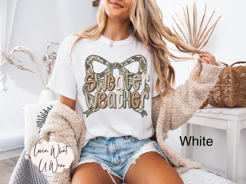 May include: White t-shirt with the text "Sweater Weather" in a bow design. The bow has a camouflage pattern. The shirt is paired with denim shorts and a beige cardigan. The words "Lovin What U Wear" are on the bottom left.