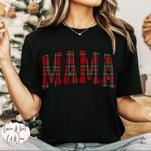 May include: Black t-shirt with the word "MAMA" in large, bold letters across the chest. The letters are filled with a red, green, and black plaid pattern. The shirt is short-sleeved.