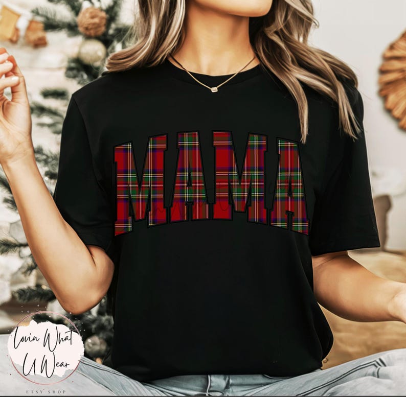 May include: Black t-shirt with the word "MAMA" in large, bold letters across the chest. The letters are filled with a red, green, and black plaid pattern. The shirt is short-sleeved.