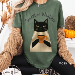 May include: A moss green t-shirt with the words "Sweater Weather" above a black cat illustration holding a coffee mug. The cat is wearing a striped sweater. The text "Moss" is visible on the bottom right.
