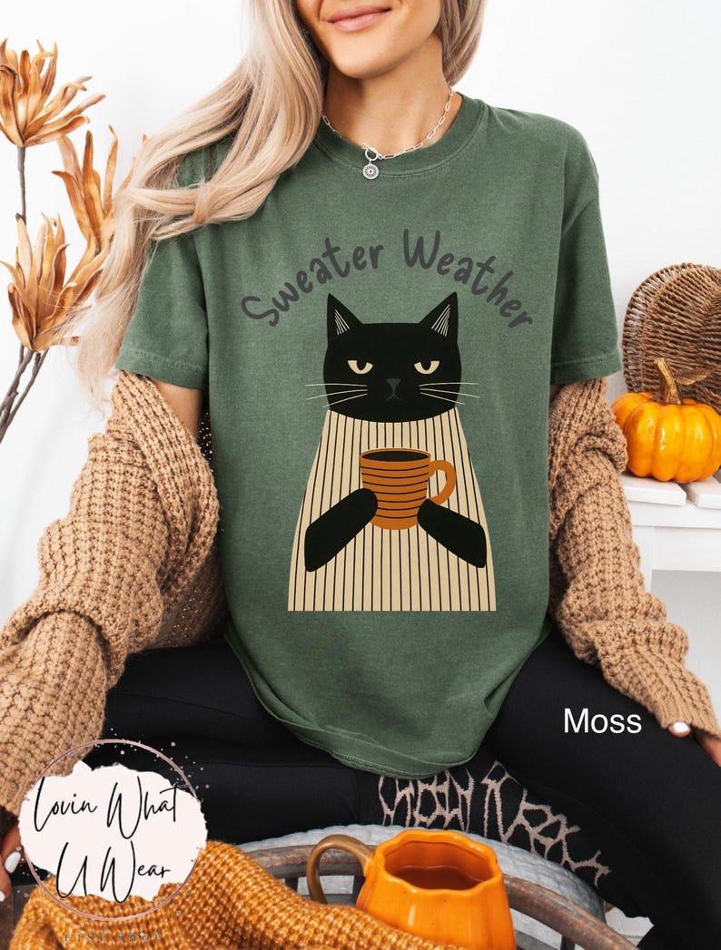 May include: A moss green t-shirt with the words "Sweater Weather" above a black cat illustration holding a coffee mug. The cat is wearing a striped sweater. The text "Moss" is visible on the bottom right.