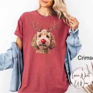 K&ouml;nnte beinhalten: Ein T-Shirt in Rotbraun mit einem Aufdruck eines braunen Hundes, der eine rote Nase und Rentiergeweih tr&auml;gt. Der Text "Lovin What U Wear" befindet sich in der rechten unteren Ecke des Shirts.