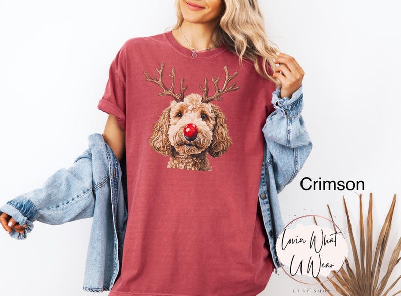 K&ouml;nnte beinhalten: Ein T-Shirt in Rotbraun mit einem Aufdruck eines braunen Hundes, der eine rote Nase und Rentiergeweih tr&auml;gt. Der Text "Lovin What U Wear" befindet sich in der rechten unteren Ecke des Shirts.