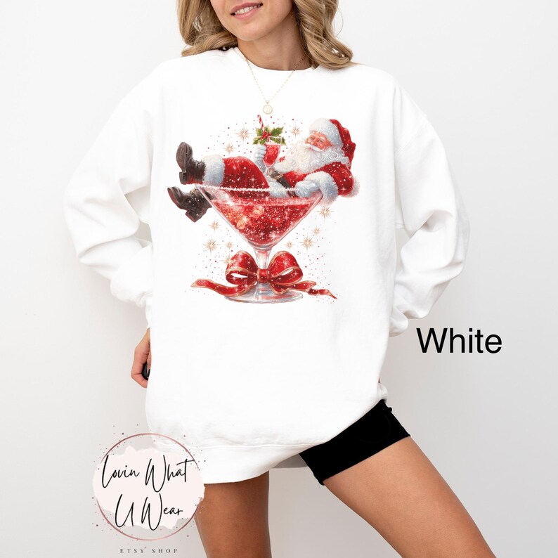 May include: White sweatshirt featuring a festive graphic of Santa Claus relaxing in a red martini glass. The design includes a red bow and the word "White". A fun, holiday-themed apparel item.