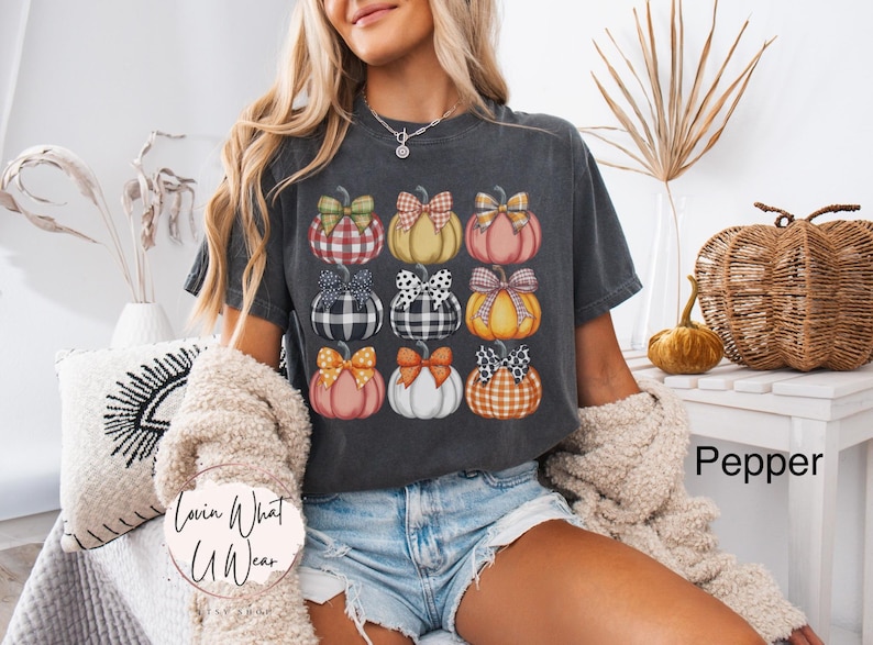 Peut inclure: T-shirt gris fonc&eacute; avec une grille de neuf citrouilles aux motifs et n&oelig;uds vari&eacute;s. Les citrouilles sont orange, rose, jaune et blanc. Le texte "Lovin What U Wear" est visible. Le t-shirt est &eacute;tiquet&eacute; "Pepper".