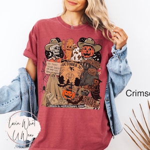 May include: Crimson-colored t-shirt with a Halloween-themed graphic. The design features a skull, a pumpkin, and other spooky characters. Text includes "October 31" and "Halloween Never Die". The shirt is a comfortable fit.