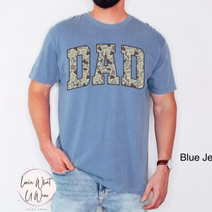 May include: A blue jean-colored t-shirt with the word "DAD" in a camouflage pattern across the chest. The shirt is a short-sleeved, crew neck style. The text "Blue Jean" is in the bottom right corner.