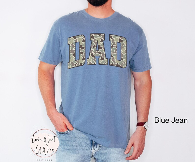 May include: A blue jean-colored t-shirt with the word "DAD" in a camouflage pattern across the chest. The shirt is a short-sleeved, crew neck style. The text "Blue Jean" is in the bottom right corner.