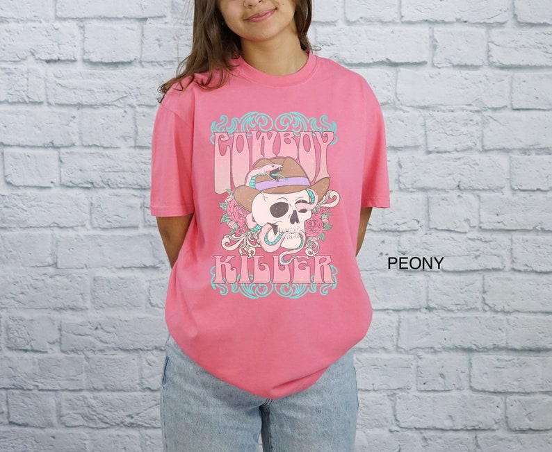 Op de afbeelding: Roze T-shirt met een afbeelding van een schedel met een cowboyhoed en een slang. De tekst "COWBOY KILLER" is onder de schedel gedrukt.