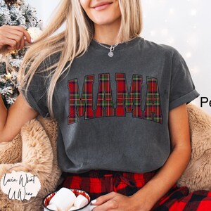 May include: A dark gray t-shirt with the word "MAMA" in a red and green plaid pattern. The shirt is part of a casual outfit, paired with red plaid pajama pants. The word "Pepper" is written in black on the right side of the image.