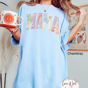 Puede incluir: Camiseta azul claro con la palabra "MAMA" en letras de patchwork. Las letras son de varios colores pastel y patrones. La camiseta es de corte holgado. Una taza con un dise&ntilde;o de sol se sostiene en la mano izquierda.