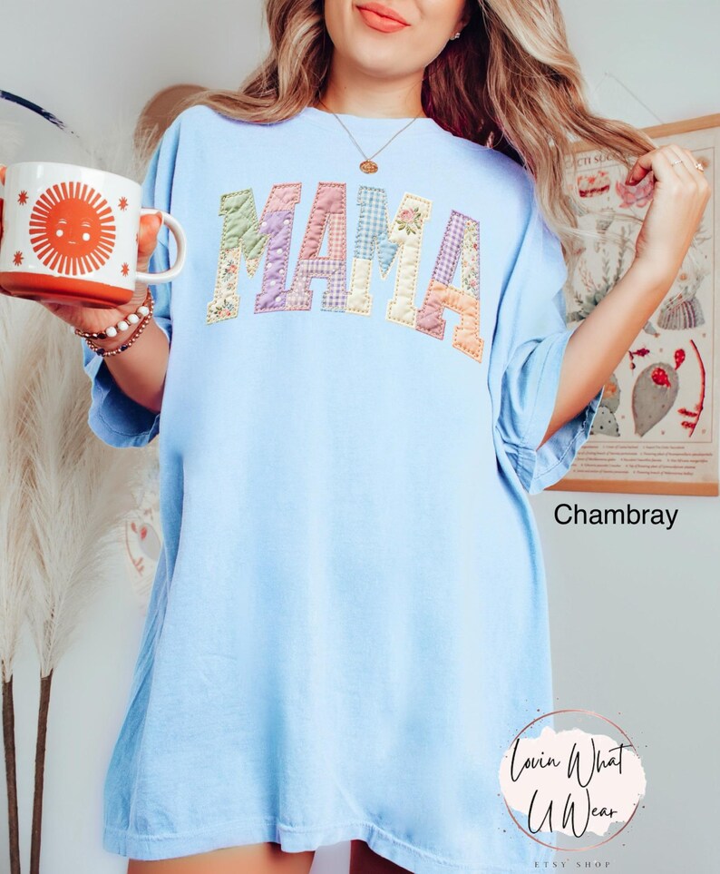 Puede incluir: Camiseta azul claro con la palabra "MAMA" en letras de patchwork. Las letras son de varios colores pastel y patrones. La camiseta es de corte holgado. Una taza con un dise&ntilde;o de sol se sostiene en la mano izquierda.