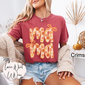 May include: Crimson-colored t-shirt with the word "Mama" in a fall-themed design. The letters are filled with pumpkins and adorned with orange bows. The shirt is paired with denim shorts.