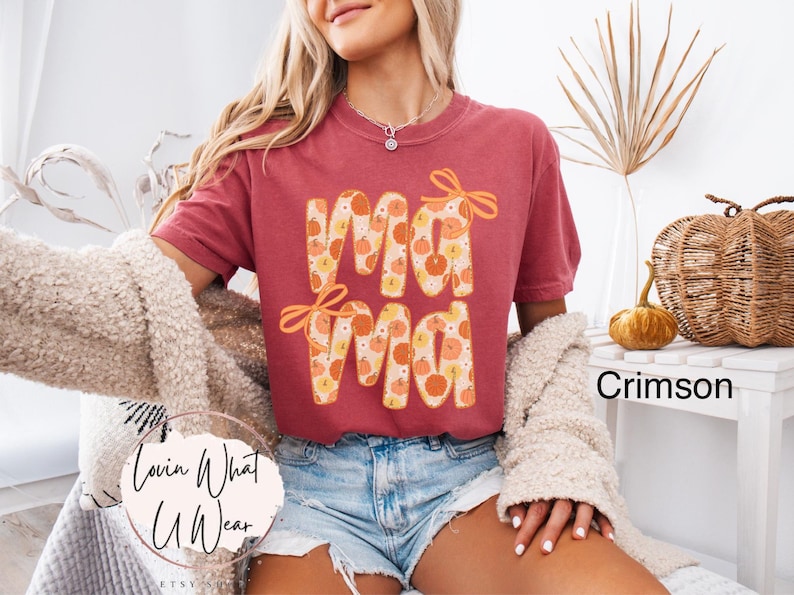 May include: Crimson-colored t-shirt with the word "Mama" in a fall-themed design. The letters are filled with pumpkins and adorned with orange bows. The shirt is paired with denim shorts.