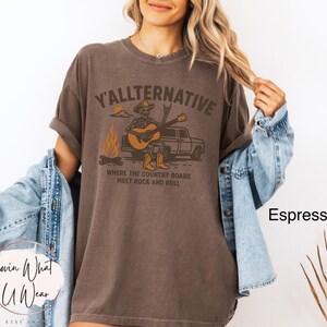 May include: Espresso-colored t-shirt with the text "Y'allternative" and a graphic of a skeleton playing a guitar. The design includes a campfire, a truck, and the phrase "Where the country roads meet rock and roll."