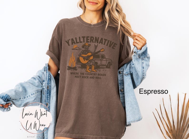May include: Espresso-colored t-shirt with the text "Y'allternative" and a graphic of a skeleton playing a guitar. The design includes a campfire, a truck, and the phrase "Where the country roads meet rock and roll."