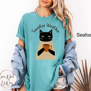 May include: Seafoam green t-shirt with a black cat graphic holding a coffee mug. The cat is wearing a striped sweater. The words "Sweater Weather" are arched above the cat. The shirt is a relaxed fit.