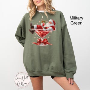 May include: Military green sweatshirt featuring a graphic of Santa Claus relaxing in a martini glass filled with red glitter. Santa is wearing a red suit and hat. A red bow adorns the base of the glass. The text "Lovin What U Wear" is at the bottom.