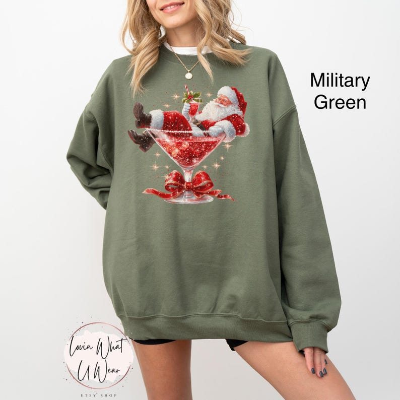 May include: Military green sweatshirt featuring a graphic of Santa Claus relaxing in a martini glass filled with red glitter. Santa is wearing a red suit and hat. A red bow adorns the base of the glass. The text "Lovin What U Wear" is at the bottom.