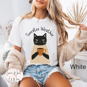 May include: White t-shirt with a black cat graphic holding a coffee mug. The text "Sweater Weather" is above the cat. The shirt is paired with denim shorts. A beige sweater is draped over the shoulders.