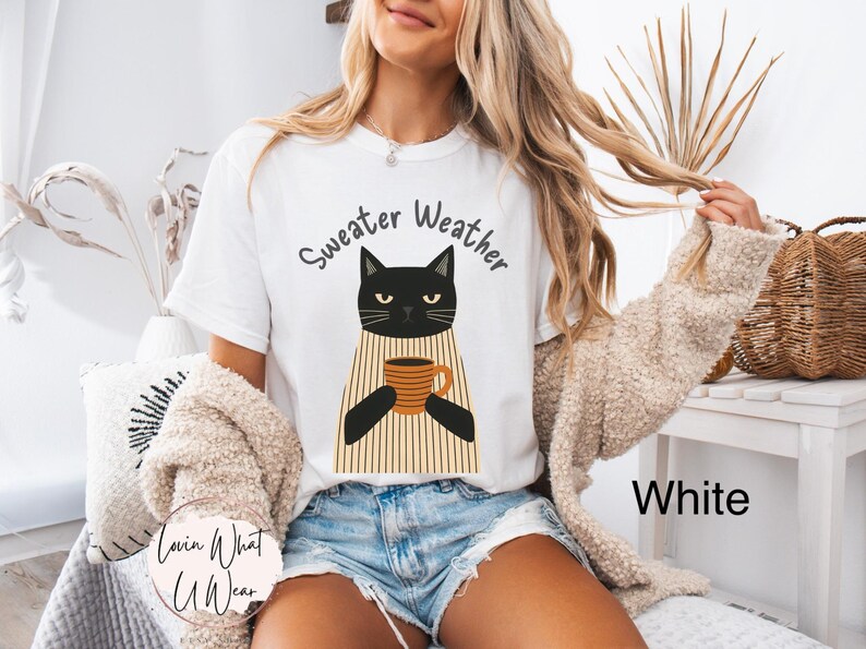 May include: White t-shirt with a black cat graphic holding a coffee mug. The text "Sweater Weather" is above the cat. The shirt is paired with denim shorts. A beige sweater is draped over the shoulders.