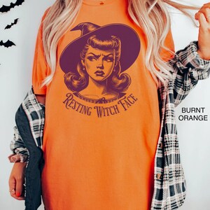 May include: A burnt orange t-shirt with a vintage illustration of a witch with a purple hat and the text "Resting Witch Face".