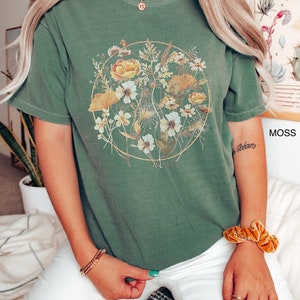 May include: A green t-shirt featuring a floral print within a circle. The flowers are yellow, white, and green. The shirt has a relaxed fit and short sleeves.