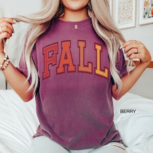 May include: A purple t-shirt with the word "FALL" printed in a retro style with orange, yellow, and red colors.