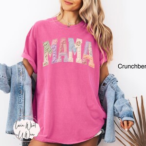 Puede incluir: Una camiseta magenta con la palabra "MAMA" en un dise&ntilde;o de patchwork en el pecho. La camiseta tiene mangas cortas y un corte holgado. El texto "Crunchberry" es visible a la derecha. El logo "Lovin What U Wear" est&aacute; abajo a la izquierda.