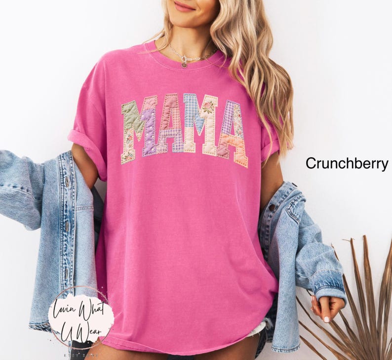Puede incluir: Una camiseta magenta con la palabra "MAMA" en un dise&ntilde;o de patchwork en el pecho. La camiseta tiene mangas cortas y un corte holgado. El texto "Crunchberry" es visible a la derecha. El logo "Lovin What U Wear" est&aacute; abajo a la izquierda.