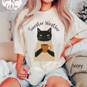 May include: Ivory-colored t-shirt with a black cat graphic holding a coffee mug. The text "Sweater Weather" is above the cat. The shirt is a relaxed fit, perfect for casual wear.