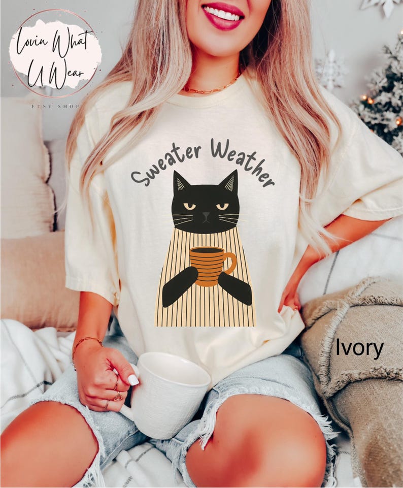 May include: Ivory-colored t-shirt with a black cat graphic holding a coffee mug. The text "Sweater Weather" is above the cat. The shirt is a relaxed fit, perfect for casual wear.
