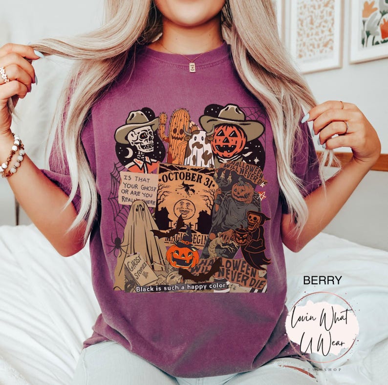May include: Berry-colored t-shirt featuring a Halloween-themed graphic. The design includes a skeleton, pumpkin, ghost, and other spooky elements. Text includes "October 31" and "Halloween Never Die". The shirt also has the text "Black is such a happy color".