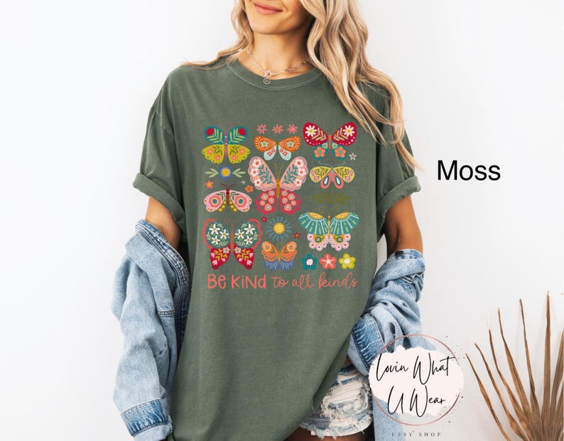 May include: A moss green t-shirt with a colorful butterfly design and the text "Be Kind to all kinds".