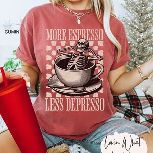 May include: A red t-shirt with the text "More Espresso Less Depresso" and a skeleton sitting in a coffee cup. The skeleton is wearing a relaxed expression and has its arms crossed. The text is in a vintage font.