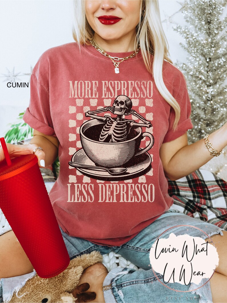 May include: A red t-shirt with the text "More Espresso Less Depresso" and a skeleton sitting in a coffee cup. The skeleton is wearing a relaxed expression and has its arms crossed. The text is in a vintage font.