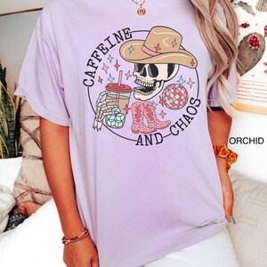 Pu&ograve; includere: Maglietta viola con una grafica di uno scheletro che indossa un cappello da cowboy, tiene una tazza di caff&egrave; e una palla da discoteca. Il testo sulla maglietta recita "Caffeine and Chaos".