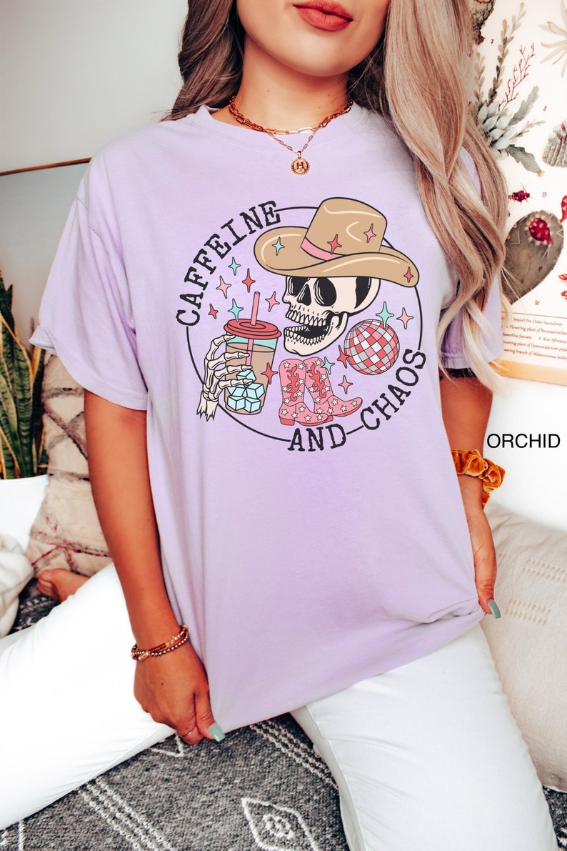 Pu&ograve; includere: Maglietta viola con una grafica di uno scheletro che indossa un cappello da cowboy, tiene una tazza di caff&egrave; e una palla da discoteca. Il testo sulla maglietta recita "Caffeine and Chaos".