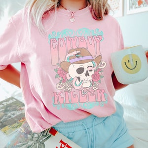 Op de afbeelding: Roze T-shirt met een grafische print van een schedel die een cowboyhoed draagt ​​met een slang eromheen gewikkeld. De tekst "Cowboy Killer" staat op het T-shirt gedrukt.