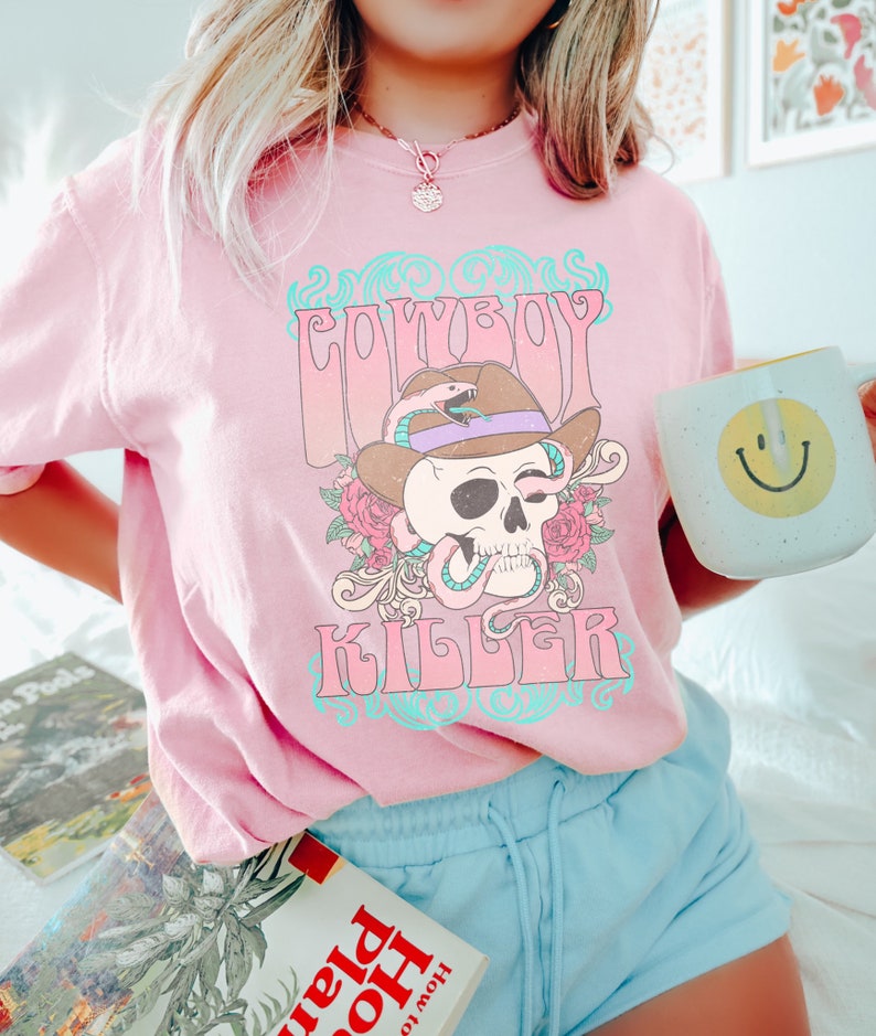 Op de afbeelding: Roze T-shirt met een grafische print van een schedel die een cowboyhoed draagt ​​met een slang eromheen gewikkeld. De tekst "Cowboy Killer" staat op het T-shirt gedrukt.