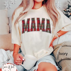 May include: Ivory-colored t-shirt with the word "MAMA" in a plaid design across the chest. The shirt is a crew neck style and has short sleeves. The image also shows a white mug and the text "Lovin What U Wear".