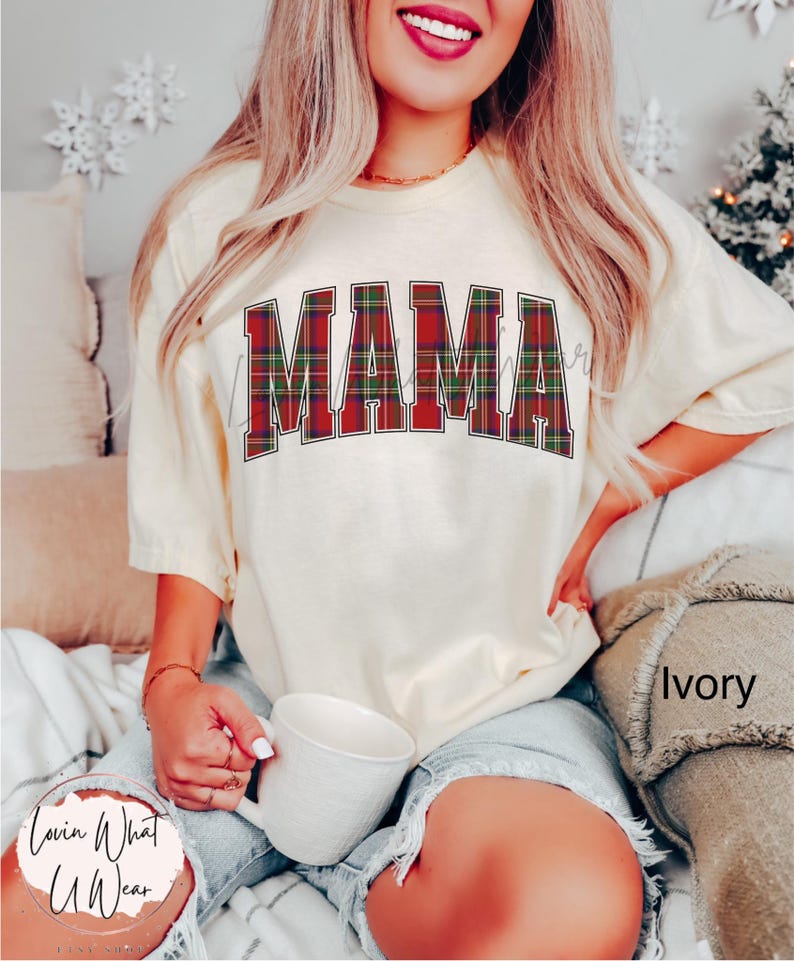 May include: Ivory-colored t-shirt with the word "MAMA" in a plaid design across the chest. The shirt is a crew neck style and has short sleeves. The image also shows a white mug and the text "Lovin What U Wear".