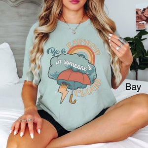 May include: A light blue t-shirt with a graphic design of a rainbow, a cloud, an umbrella, and lightning bolt. The text on the shirt reads "Be a rainbow in someone's cloud."