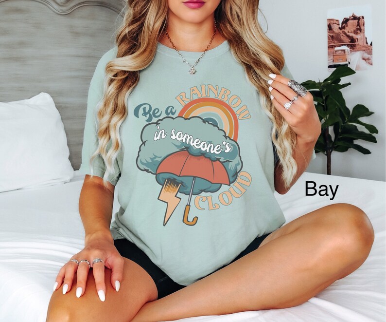 May include: A light blue t-shirt with a graphic design of a rainbow, a cloud, an umbrella, and lightning bolt. The text on the shirt reads "Be a rainbow in someone's cloud."