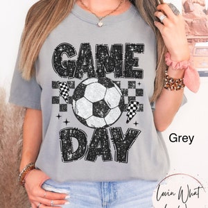 May include: Grey t-shirt with a soccer ball graphic and the words "GAME DAY" in a distressed, glittery font. The design includes checkered flags and stars. The shirt is a casual, comfortable style.