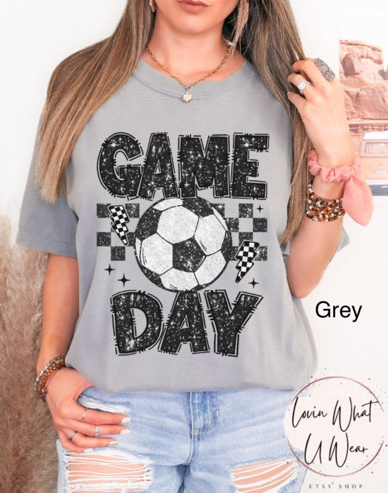 May include: Grey t-shirt with a soccer ball graphic and the words "GAME DAY" in a distressed, glittery font. The design includes checkered flags and stars. The shirt is a casual, comfortable style.