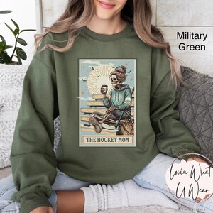 May include: A green sweatshirt featuring a graphic of a skeleton holding a hockey stick and a cup of coffee. The graphic is in a vintage style and says "The Hockey Mom".