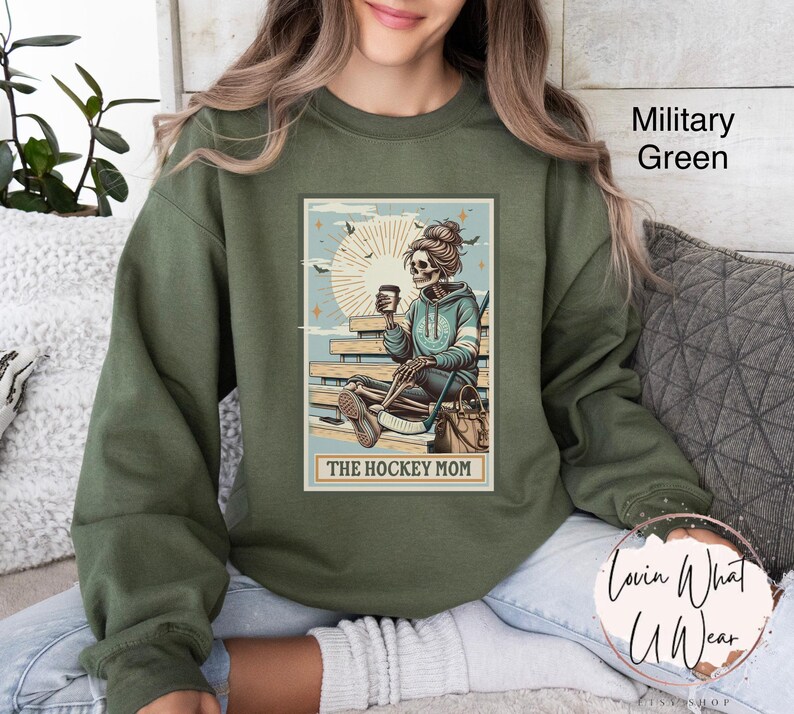 May include: A green sweatshirt featuring a graphic of a skeleton holding a hockey stick and a cup of coffee. The graphic is in a vintage style and says "The Hockey Mom".